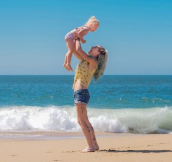 Happy mother lifting child on a sandy beach under clear blue skies.