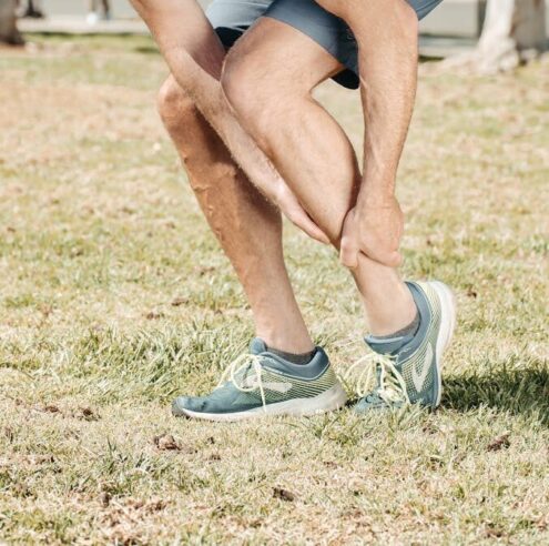 Man holding his injured ankle outdoors in a park, highlighting running-related pain or sport injury.