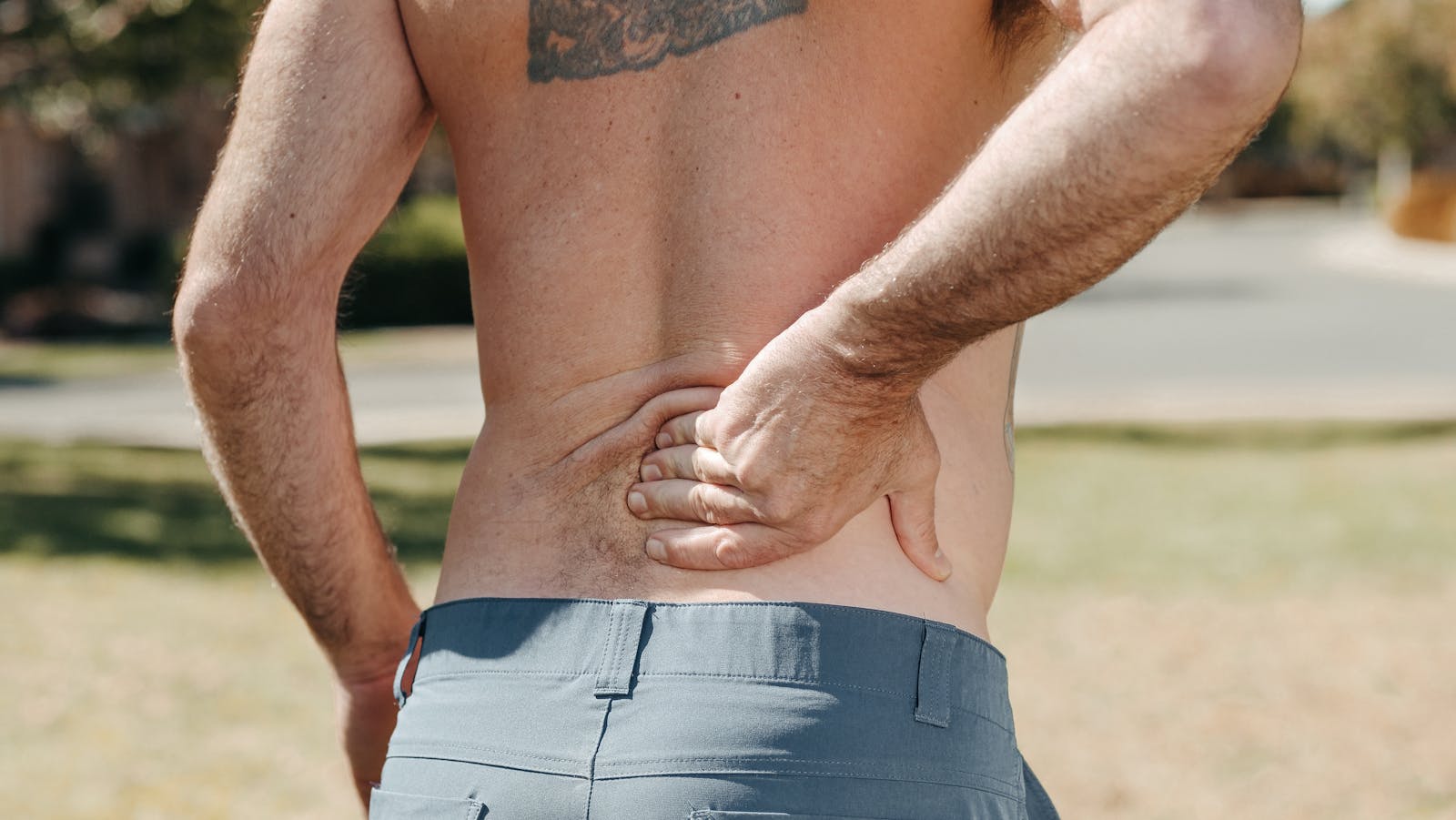 Photo by Kindel Media Shirtless man holding his lower back outdoors indicating pain or discomfort.